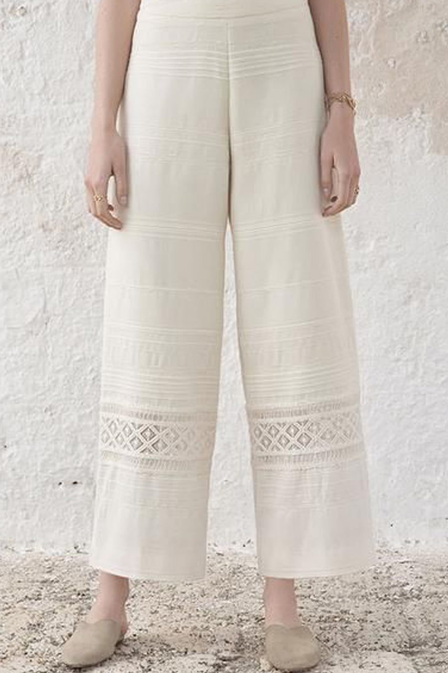 White pants with lace details worn by a person against a textured white wall.