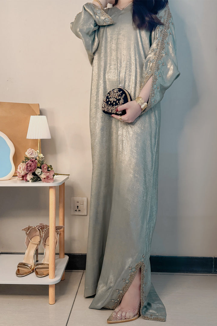Woman in a long, light-colored dress holding a clutch in a room with a side table and lamp.