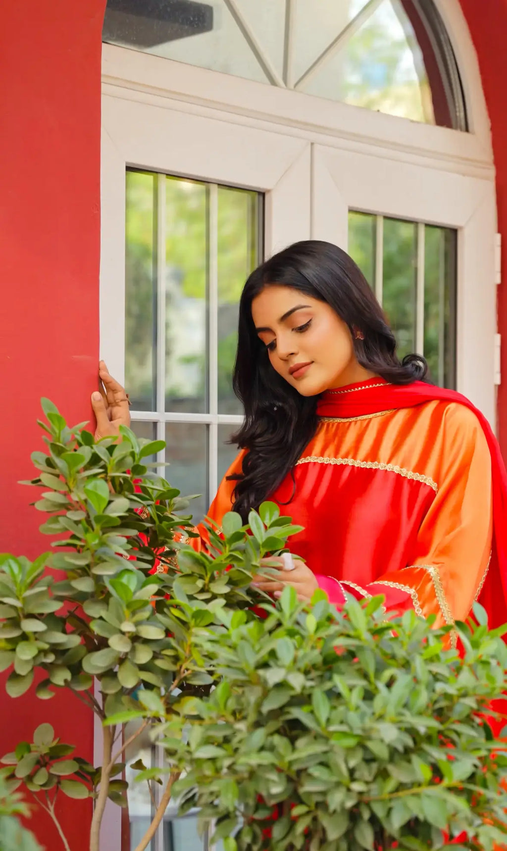 Woman in a fiery sunset glamour outfit tending to plants in front of a red wall with a window.