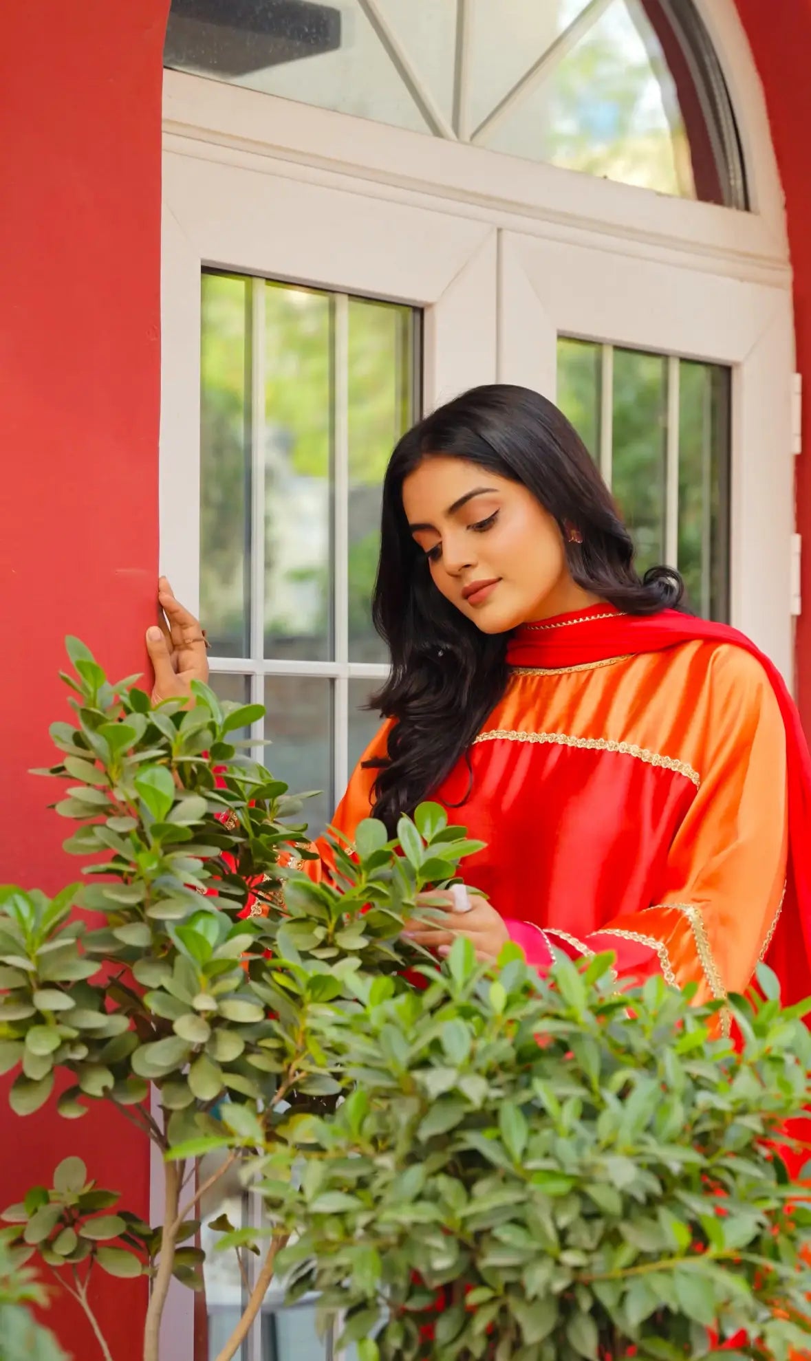 Woman in a fiery sunset glamour outfit tending to plants in front of a red wall with a window.