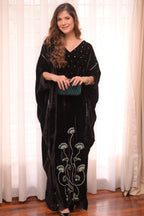 Woman wearing a black embroidered dress holding a green clutch in front of white curtains.