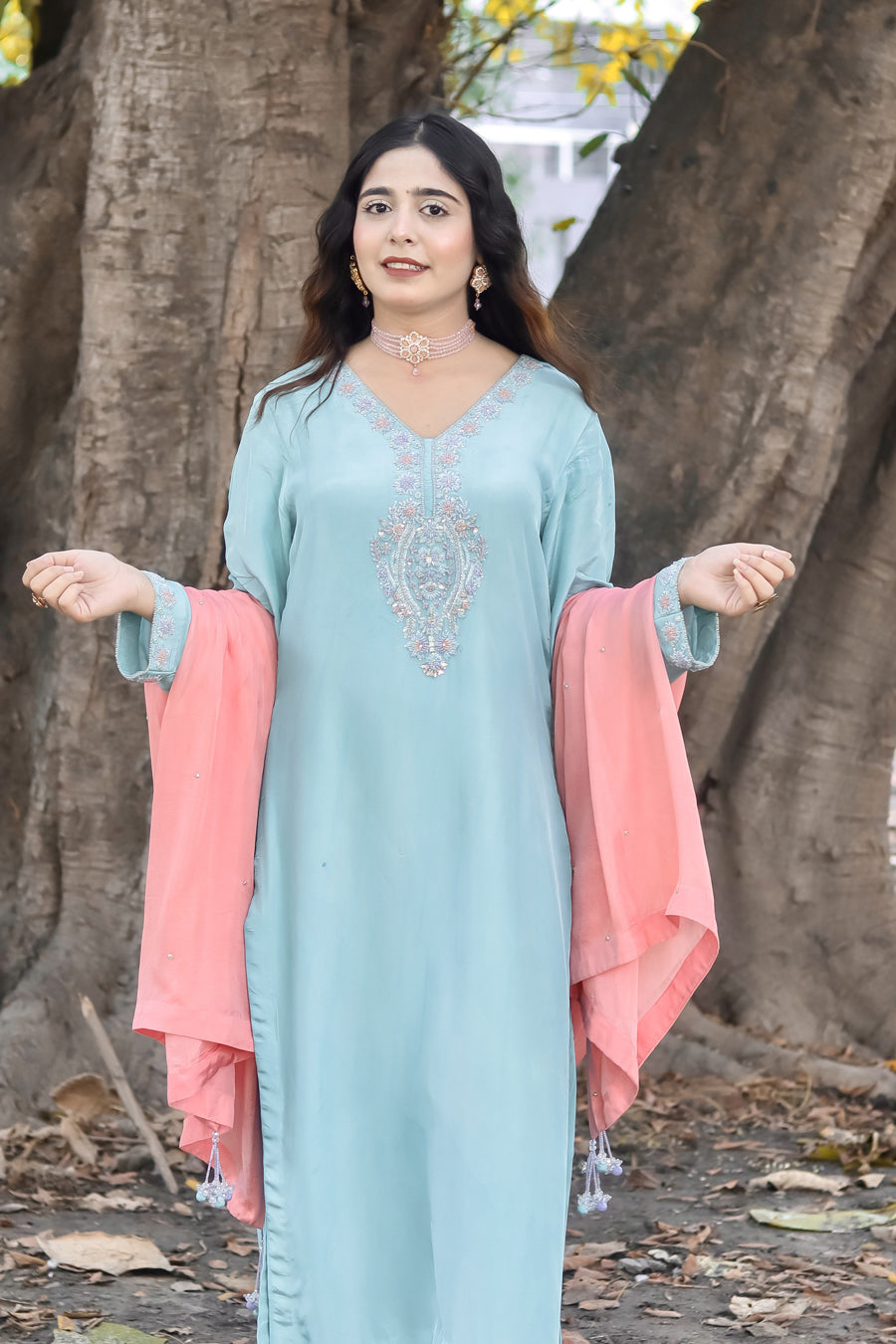 Woman in a light blue dress with pink dupatta standing outdoors near large trees.