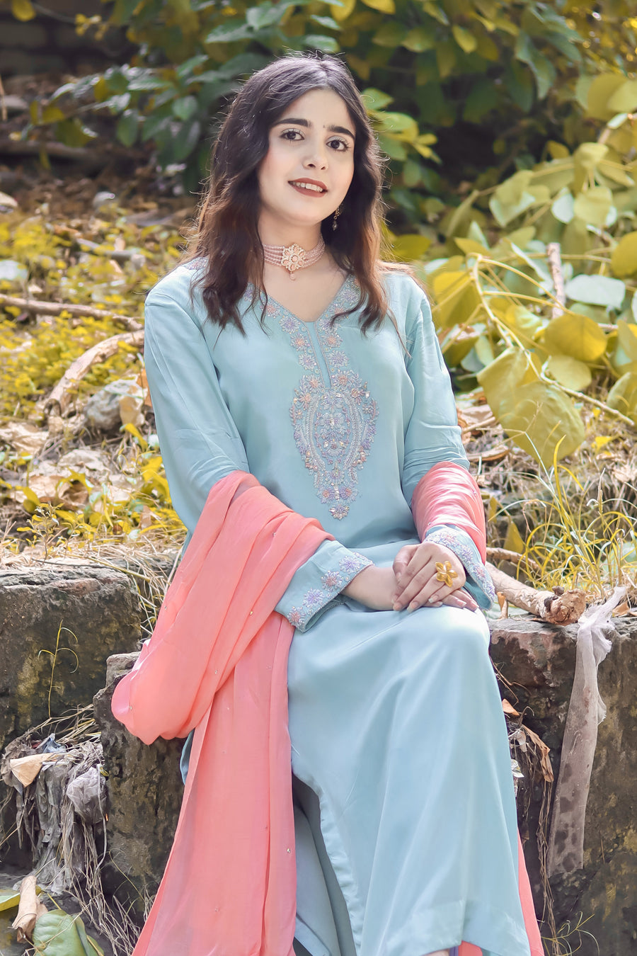 Woman in a light blue traditional outfit with a pink dupatta sitting outdoors among greenery.