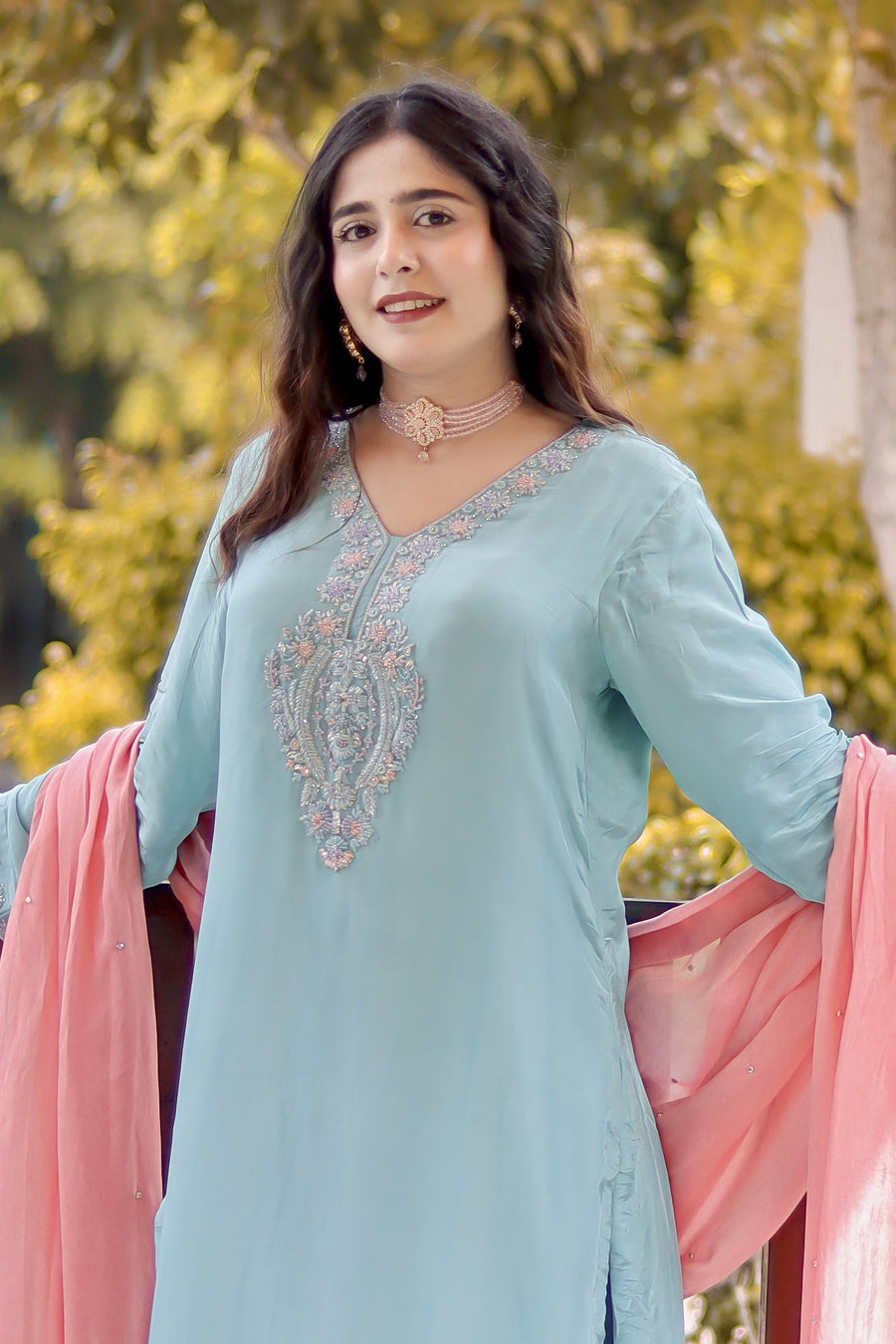 Woman wearing a light blue embroidered outfit with pink dupatta against a blurred natural background