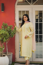 Woman in a lemon yellow base highlighted with subtle threadwork and a contrasting pastel dupatta standing in front of a red wall and glass door.