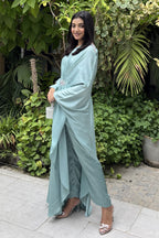Woman in a light blue traditional outfit standing in front of green plants.
