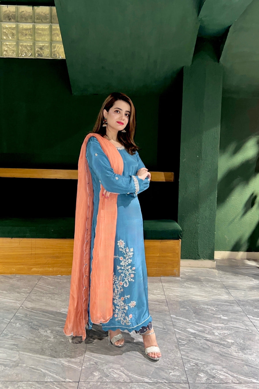 Woman in a blue traditional outfit with an orange dupatta standing indoors.