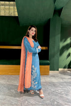 Woman in a blue traditional outfit with an orange dupatta standing indoors.